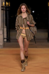 Model walking a nighttime runway in a striped olive-green coat over beige bikini bottoms, a wide tan belt, patterned boots, and a large bag.
