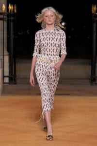 Runway model walks in a cream jumpsuit with brown teardrop print, a wide beige belt, and dangling earrings on a nighttime catwalk.