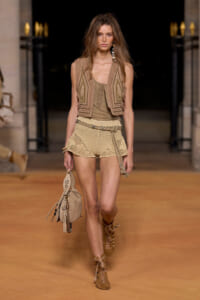 Model walking a runway in beige boho outfit with a knitted vest, crochet shorts, and lace-up sandals, carrying a tan bag.
