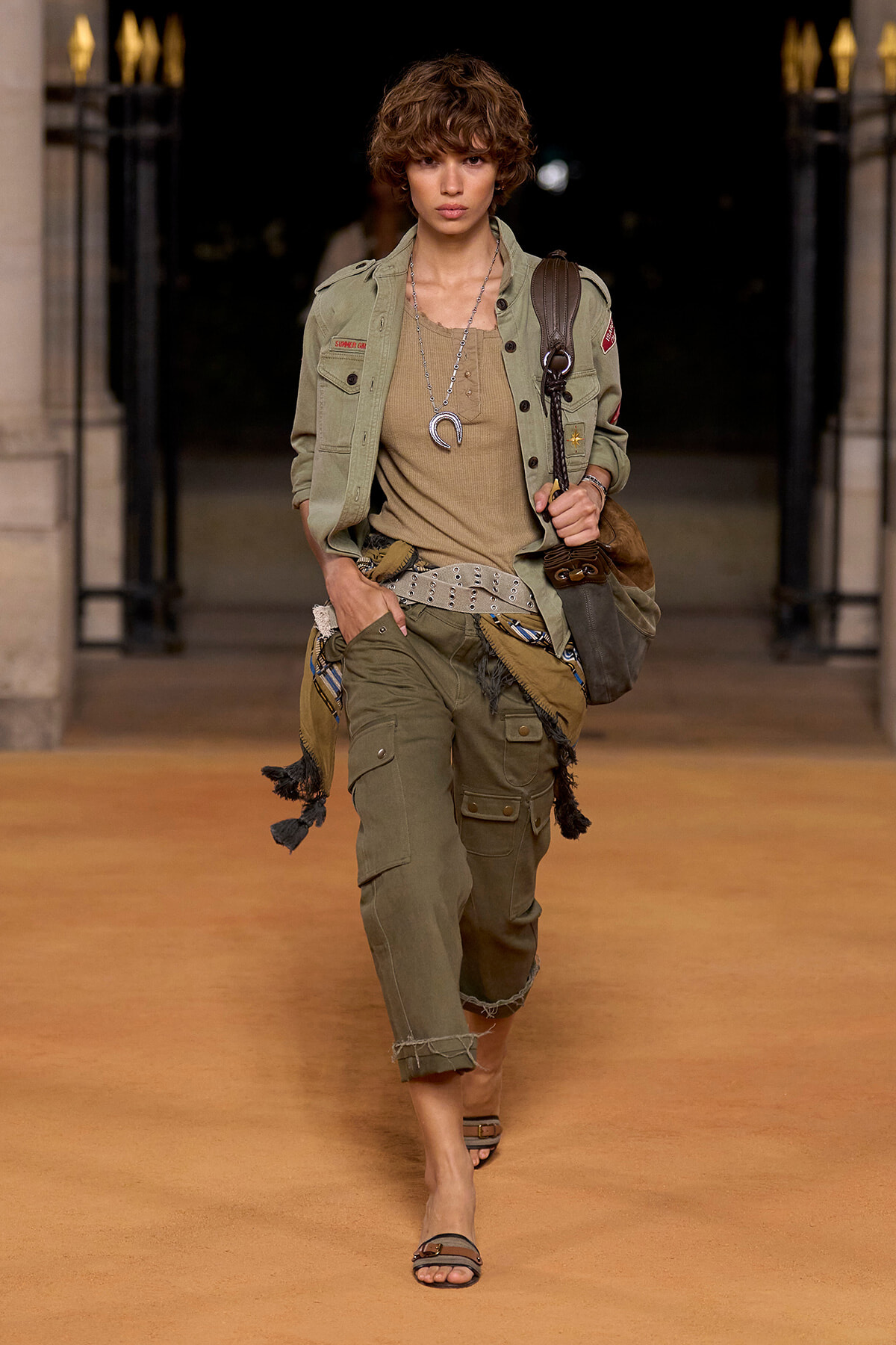 Female model walking a runway in an olive military-inspired outfit: cargo pants, beige top, belt, layered necklaces, and a brown shoulder bag.