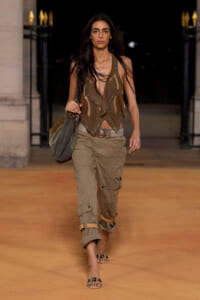 Female model walking a night runway in tan cargo pants and a studded brown vest, carrying an olive slouch bag on her shoulder.