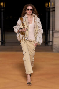 Model walking a runway in beige utility jacket, matching cargo pants, and sunglasses with a tan tote bag.