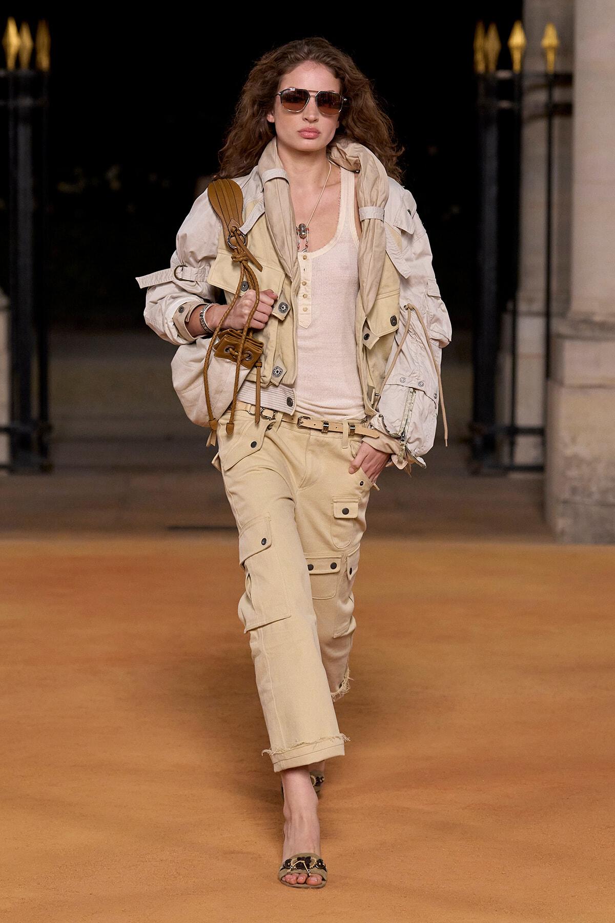 Model walking a runway in beige utility jacket, matching cargo pants, and sunglasses with a tan tote bag.