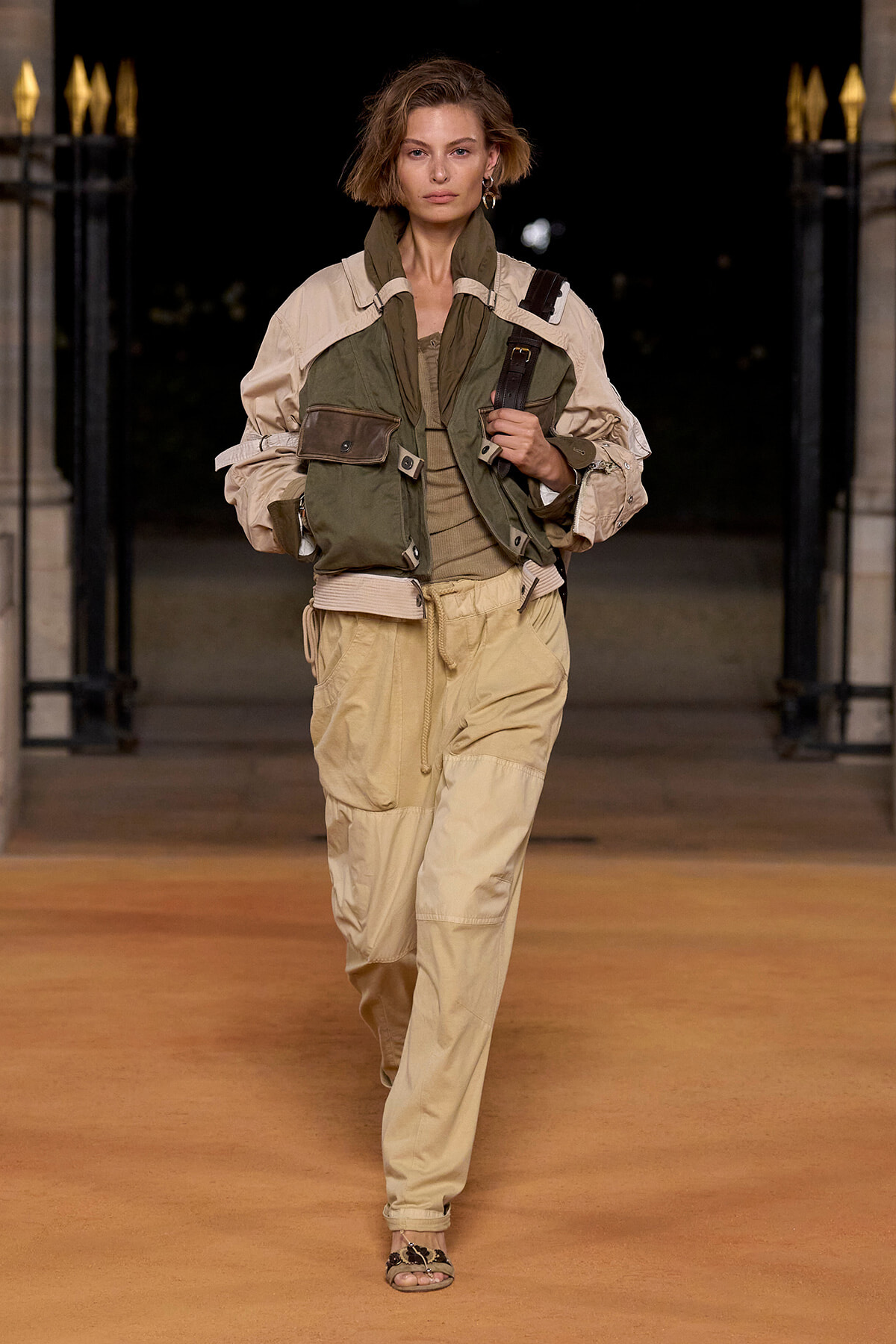 Model walking a runway at night in a beige utility jacket layered over an olive vest and tan cargo pants, holding a strap.