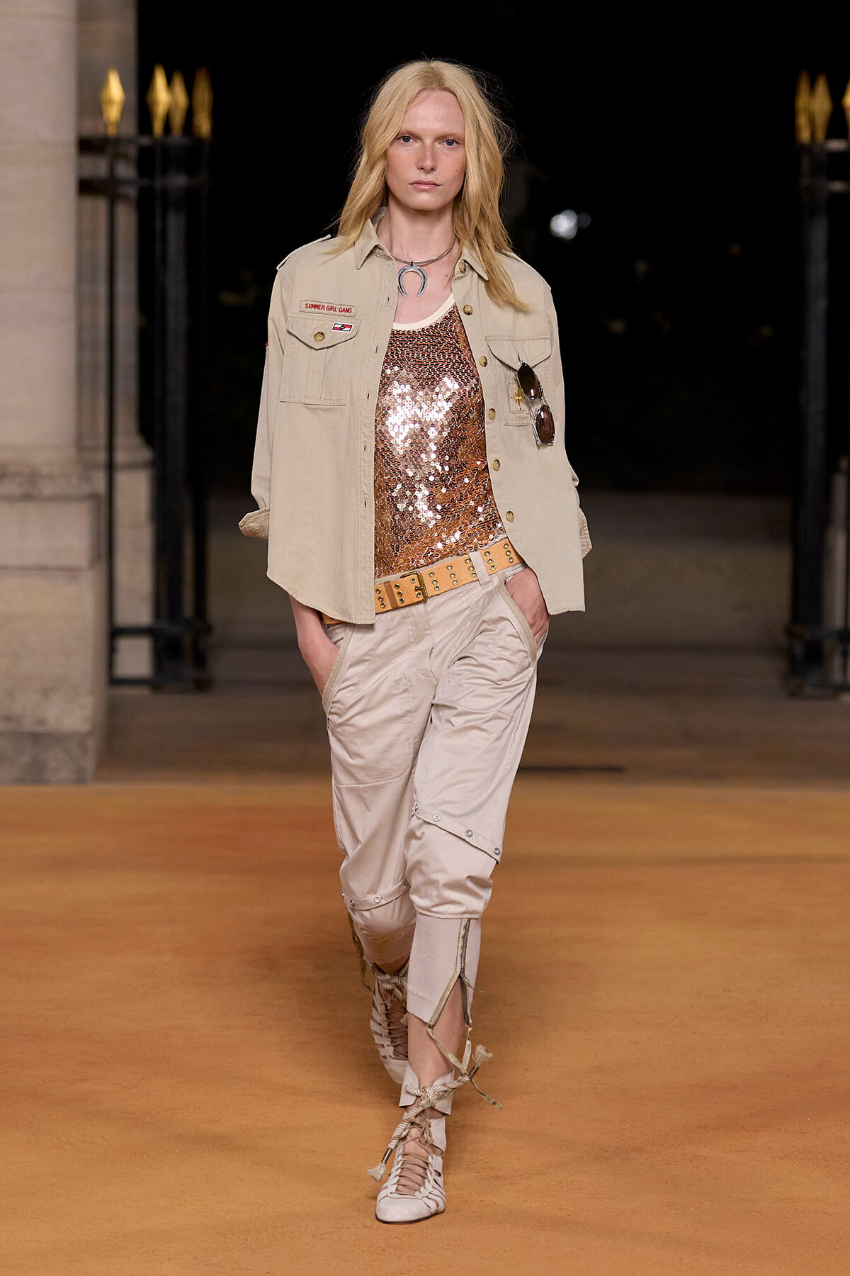 Model walking the runway in a beige safari jacket over a copper sequined top with beige cargo pants and tan lace-up boots.