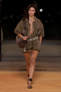 Model walking a runway in a khaki safari-inspired outfit with a loose shirt, matching shorts, wide belt, and a large patterned bag over the shoulder.