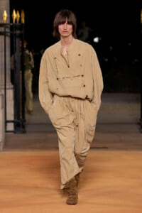Male model walking a runway in a beige oversized jumpsuit with multiple pockets and brown boots.