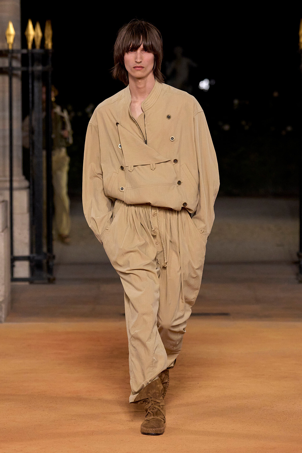 Male model walking a runway in a beige oversized jumpsuit with multiple pockets and brown boots.