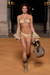 Model walking runway in a beige bandeau bikini with matching bottoms, carrying a beige jacket and a large tan bag, desert-toned boots, evening venue.