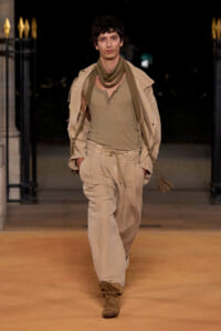 Male model walking a runway in a beige safari-inspired outfit with a tan scarf and rope belt.