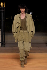 Male model walks a nighttime runway in a loose khaki utility jacket and drawstring cargo pants with tan boots under warm lighting.