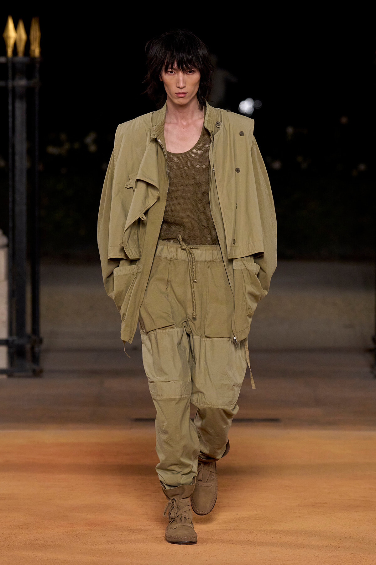 Male model walks a nighttime runway in a loose khaki utility jacket and drawstring cargo pants with tan boots under warm lighting.