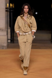 Runway model in a tan utility jumpsuit with a wide beige belt and crossbody strap.