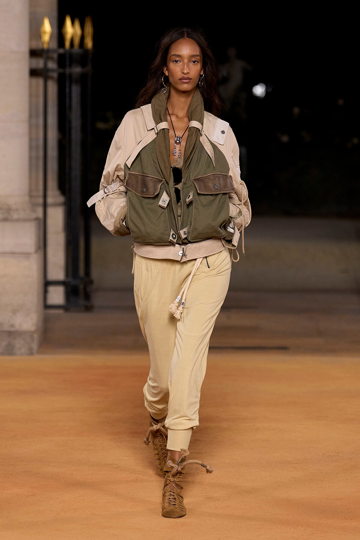 Runway model wearing a tan and olive utility jacket with cargo pockets paired with beige pants and tan lace-up boots.