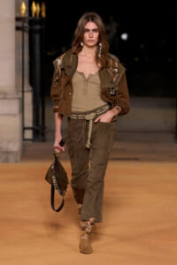 Model walking a fashion runway in olive-green top, brown jacket, and cargo pants with a rope belt and boots, carrying a brown bag.