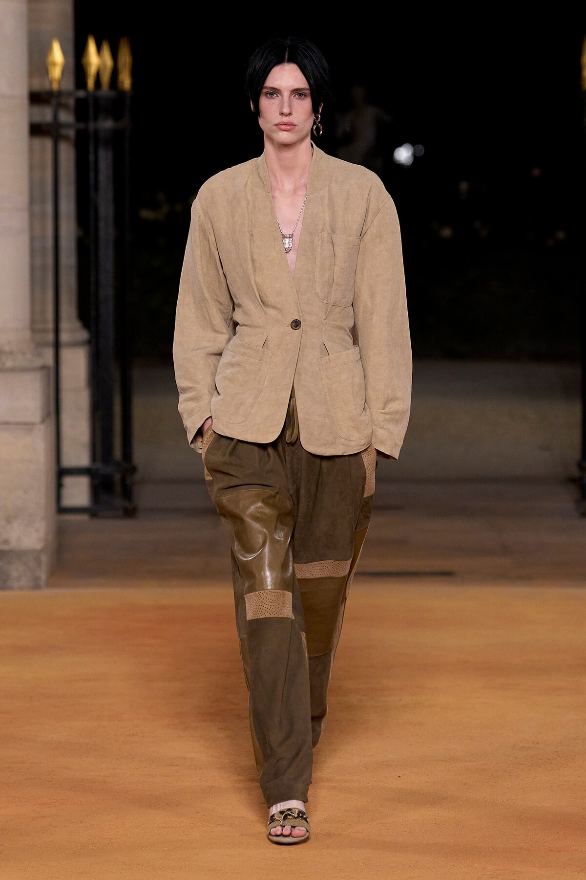 Model walking a runway at night in a beige blazer with large pockets and a single button, paired with patchwork brown pants and open-toed sandals, wearing a layered necklace.