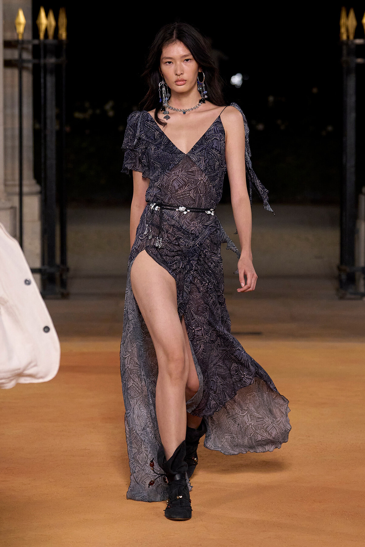 Model walking a runway at night in a sheer, dark patterned gown with a high slit, a thin decorative belt, and bold jewelry.