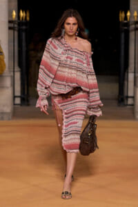 Model walking a runway in a pink striped off-shoulder knit top and matching midi skirt with ruffled edges, brown belt, and brown handbag.