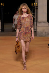 Model walking a runway in a multicolored distressed-knit mini dress, carrying a tan suede handbag and wearing lace-up sandals and statement earrings.