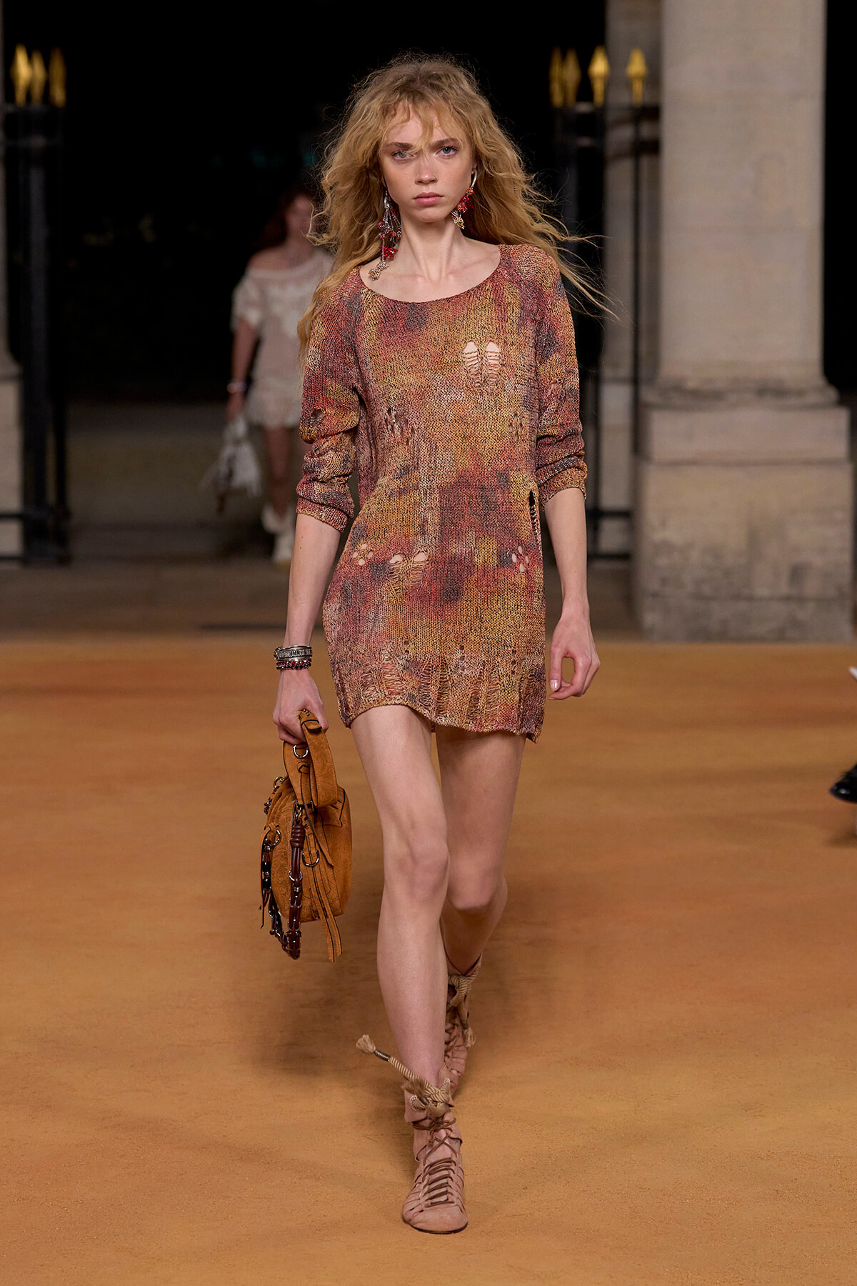 Model walking a runway in a multicolored distressed-knit mini dress, carrying a tan suede handbag and wearing lace-up sandals and statement earrings.