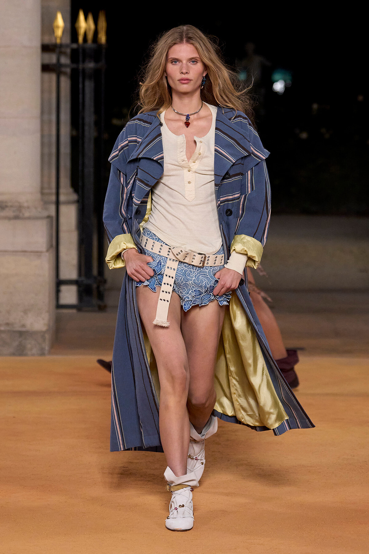 Model walking a runway in a blue striped trench coat over a cream top and blue lace shorts with a wide beige belt and white boots.