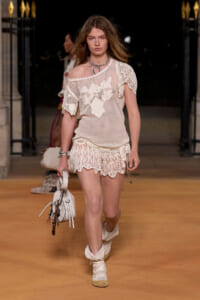 Model walks the runway in a beige crochet top with floral lace accents and matching crochet shorts, carrying a white handbag.