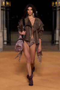 Runway model wearing a large brown patterned jacket over a bikini, burgundy boots, and a oversized patterned bag slung over the shoulder at night.