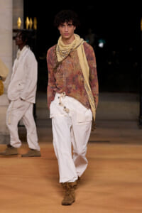 Male model walks the runway in a distressed multicolor knit sweater, beige scarf, white cargo pants, and brown boots.