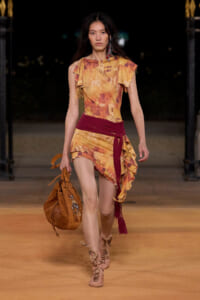 Model walking a runway at night in an orange-brown tie-dye asymmetric dress with a burgundy sash, carrying a tan leather bag.