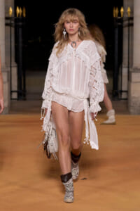 Model walking the runway in a white lace boho blouse with matching shorts, patterned boots, and a white handbag.