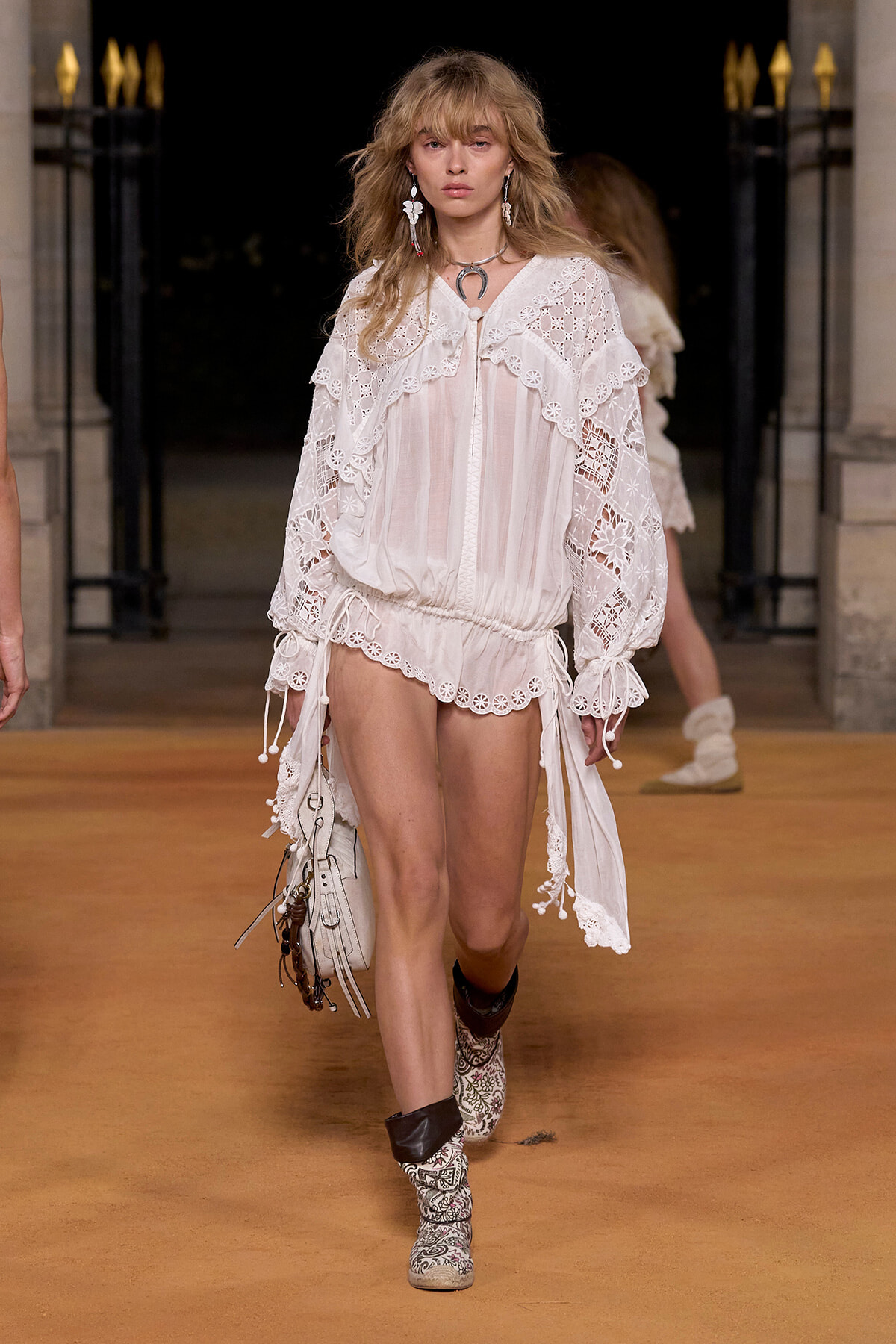 Model walking the runway in a white lace boho blouse with matching shorts, patterned boots, and a white handbag.