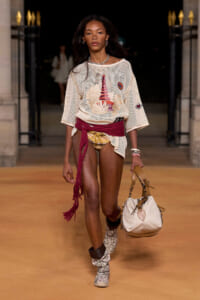 Model walks a runway in a boho-chic look: oversized beige mesh top with a graphic print, burgundy sash tied at the waist, patterned bikini bottoms, and snakeskin-effect ankle boots; carries a large beige handbag and wears layered jewelry.