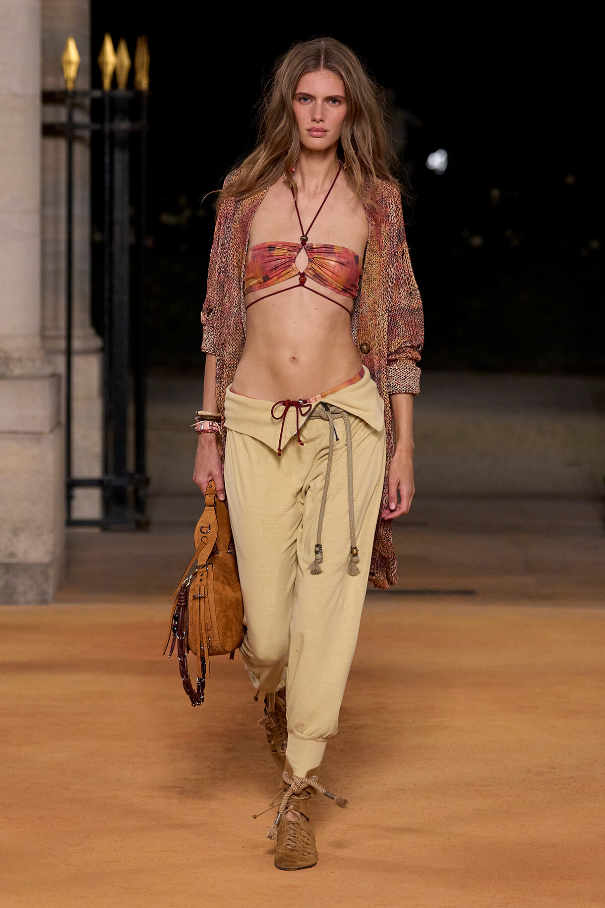 Female model walks a nighttime runway in a bohemian outfit: tie-dye bandeau top, cream drawstring pants, and a woven beige cardigan, carrying a tan suede bag.
