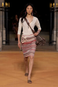 Model walking the runway in a beige long-sleeve top, striped knit pencil skirt with belt, brown crossbody bag, and flat sandals.