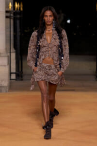 Model walks a nighttime runway wearing a brown paisley two-piece set with a cropped top and mini skirt, paired with black ankle boots and long dangling earrings.