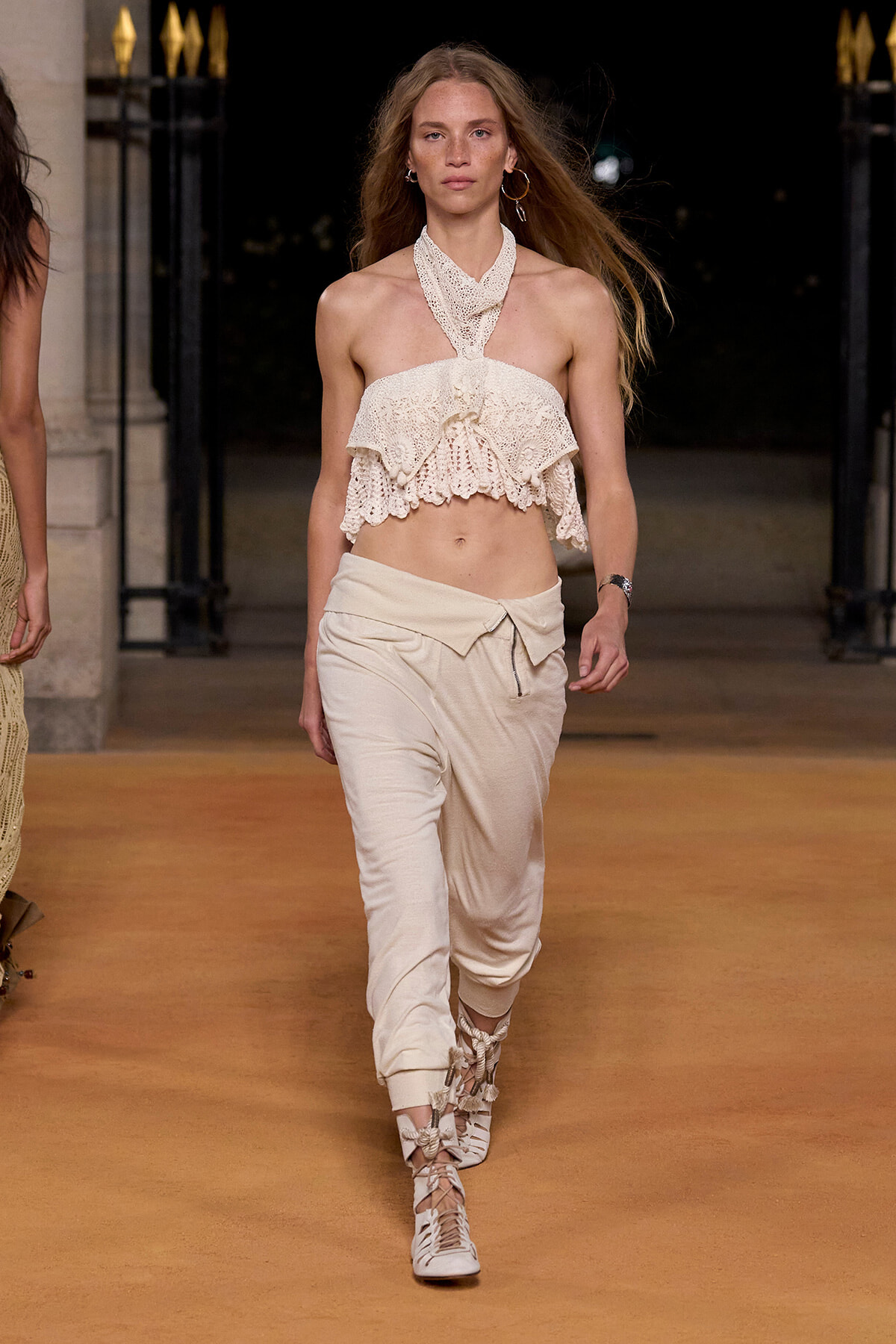 Model walking a runway in a cream crochet halter top with a ruffled peplum and beige drawstring pants, lace-up sandals visible. A fashion show look.