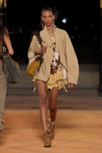 Model walking a runway in a beige oversized jacket, ruffled tan shorts, and rope-laced tan boots, carrying a tan shoulder bag with jewelry accents.