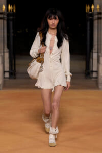 Model walking a nighttime runway in a cream-beige button-down romper, with a tan shoulder bag and light sneakers/boots.