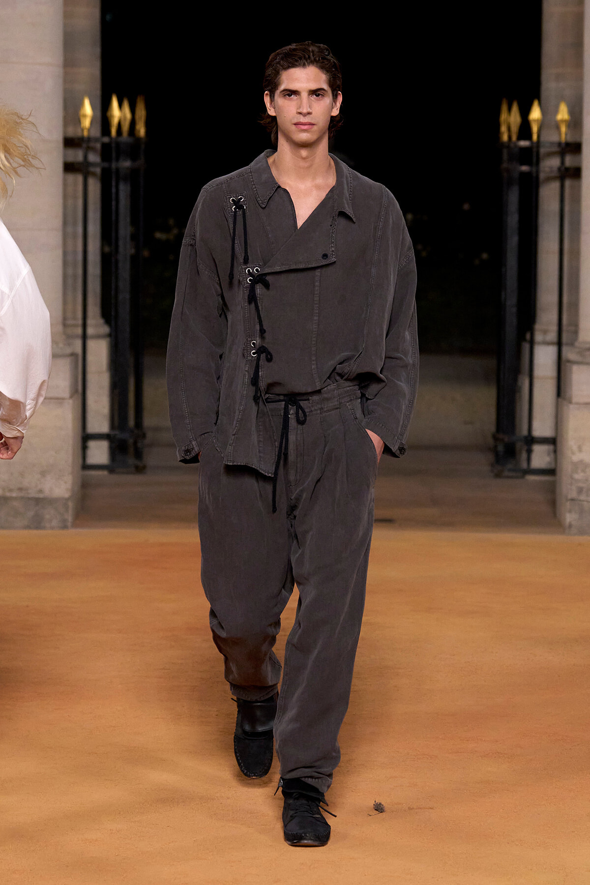 Male model walks a runway in a dark gray loungewear set with lace-up detailing on the front, black shoes.
