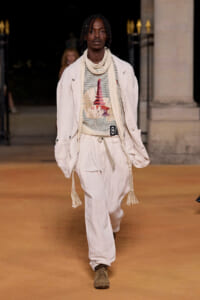 Male model walks the runway in an off-white relaxed suit with a scarf and tasseled belt, beige boots.
