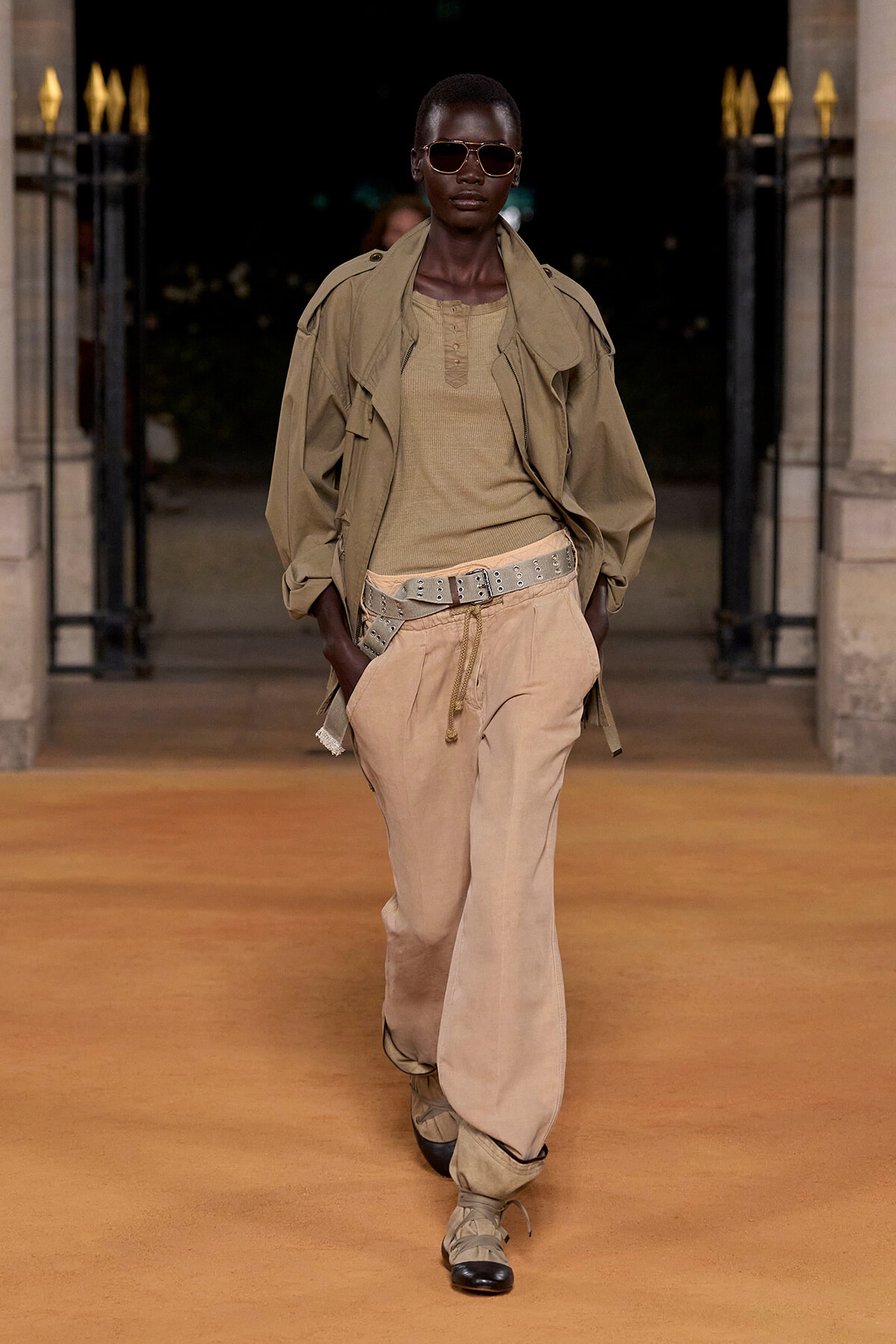 Model walking on an orange runway in a khaki trench, beige top, wide belt, and sunglasses; urban backdrop behind gates at night.