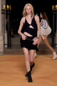 Female model walking a runway in a black cut-out lace halter dress with a fringed mini skirt, at a night show.