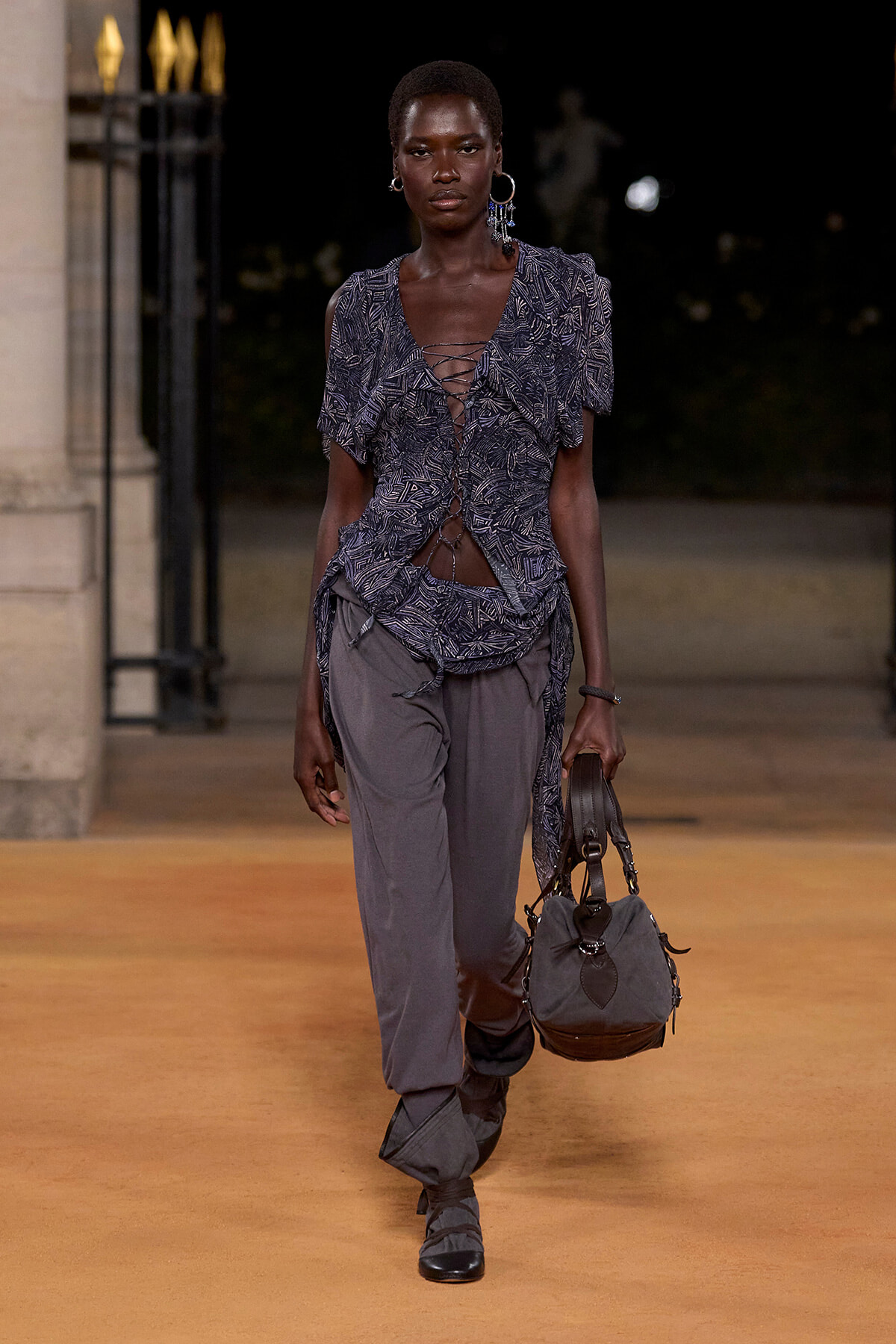 Model walks a nighttime runway in a blue patterned lace-up top, loose grey pants, and a gray handbag.