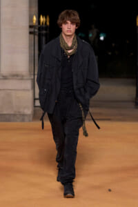 Male fashion model walking a runway at night in an all-black outfit with a olive scarf and layered jacket.