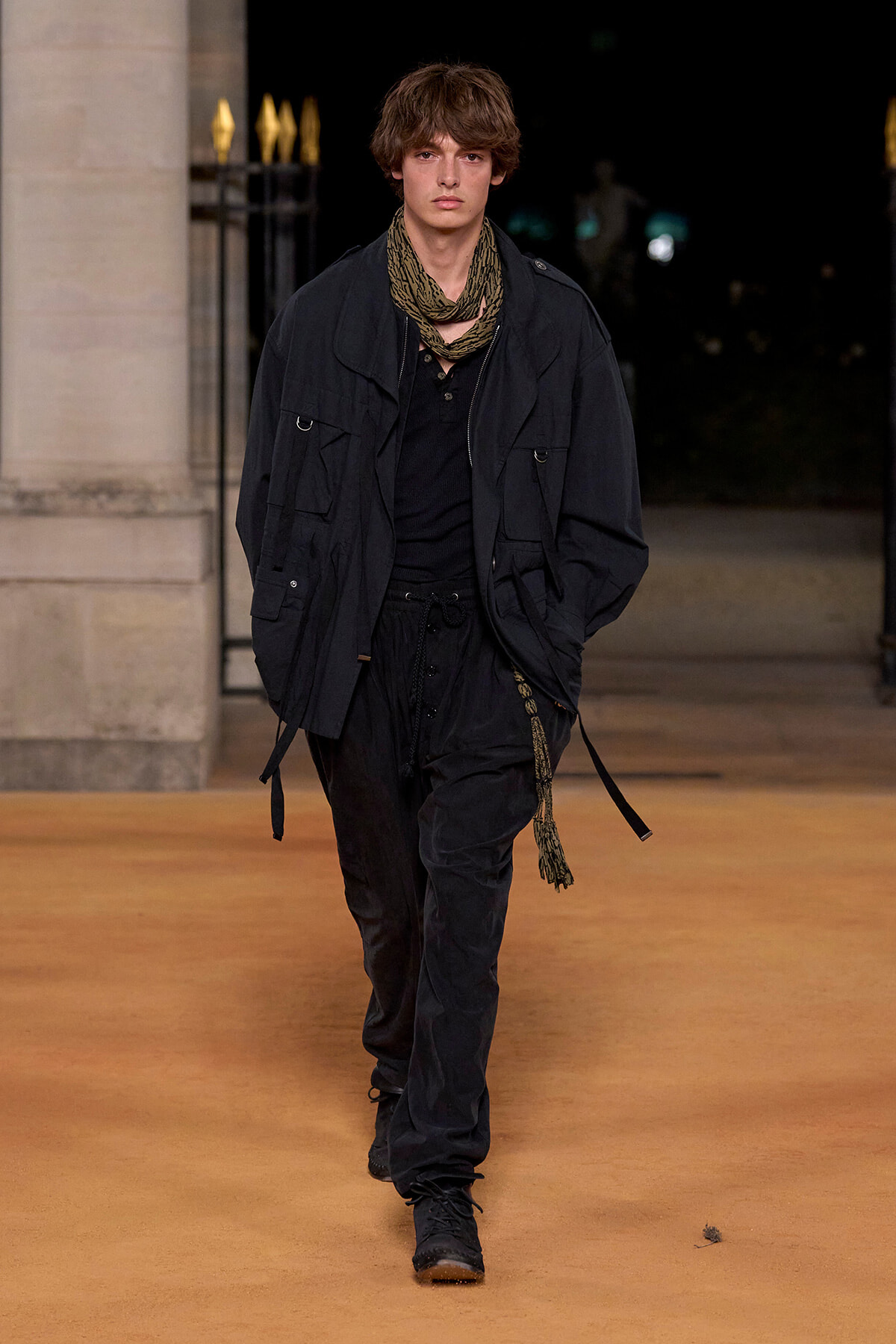 Male fashion model walking a runway at night in an all-black outfit with a olive scarf and layered jacket.