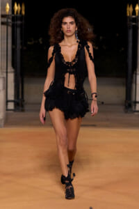 Model walking a night runway in a black lace crop top and layered mini skirt with cutouts and sandals.