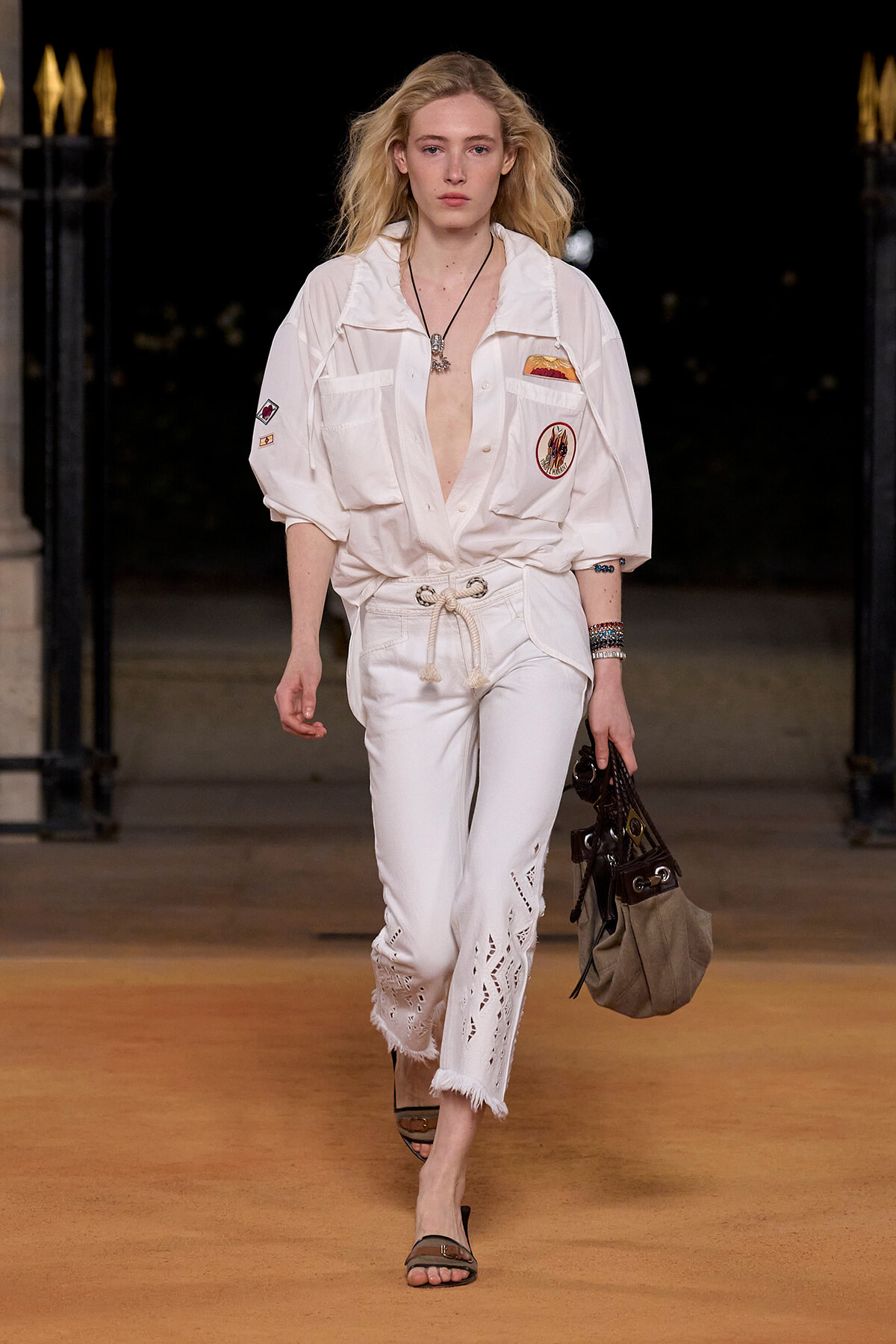 Model walking a runway in a white patchwork jumpsuit with a rope belt, carrying a beige bag, at nightfall.