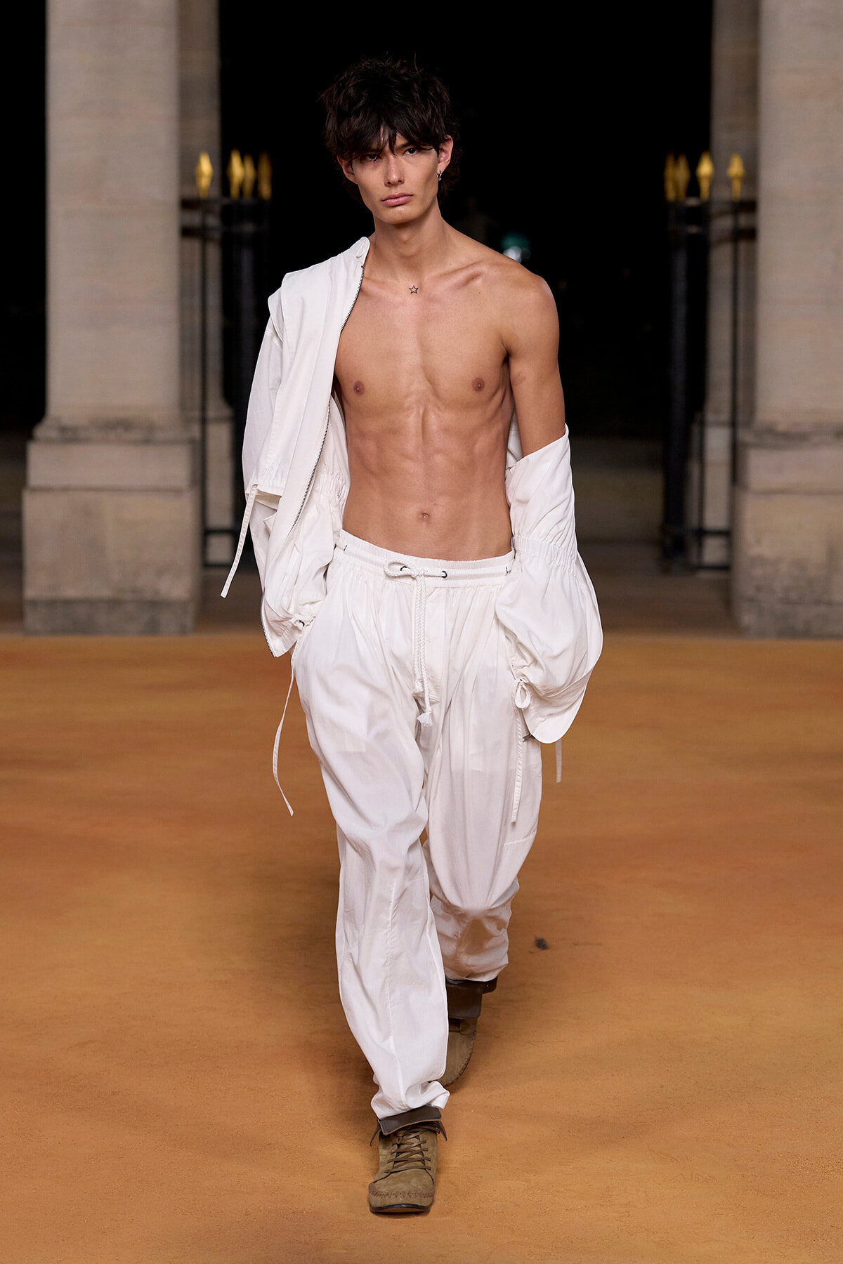 Male fashion model walking a runway at night, shirtless with white baggy pants and a white jacket draped over one shoulder, a confident pose.