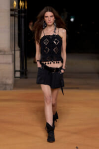 Model walking a runway in a black lace crop top with scalloped edges and black utility shorts, accessorized with long earrings.