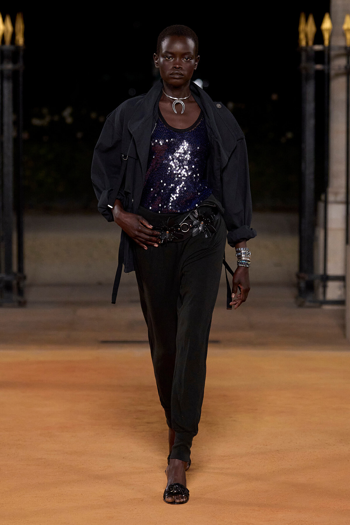 Model walking a nighttime runway in a dark oversized jacket over a blue sequined top, black pants, a wide belt, and stacked bracelets.